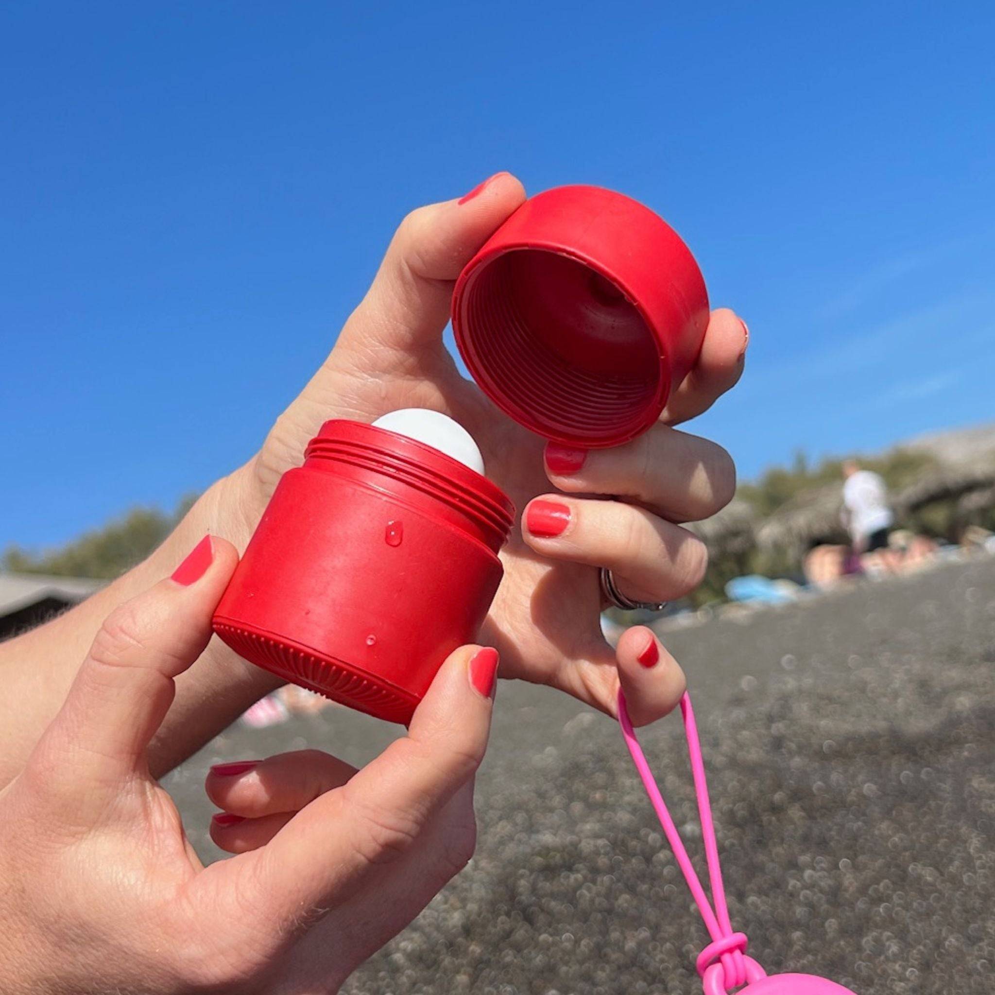 Red refillable roll-on travel applicator with lid off held in hands at a beach