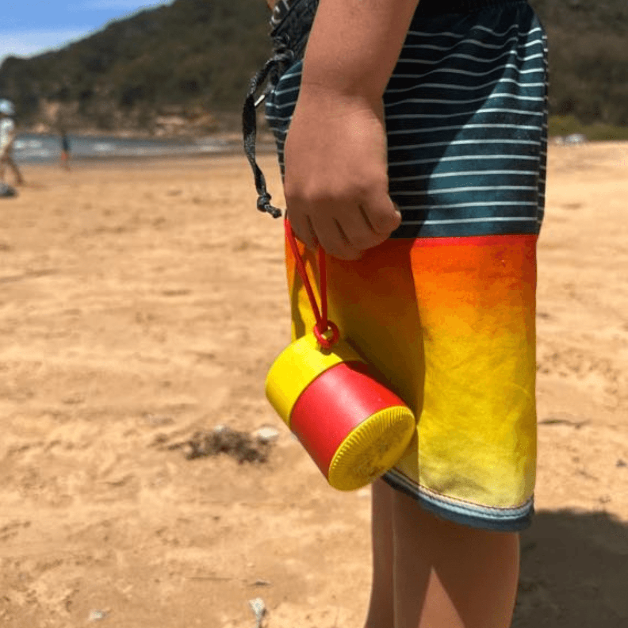 Red and yellow refillable roll-on travel applicator held by a boy in front of his shorts at the beach